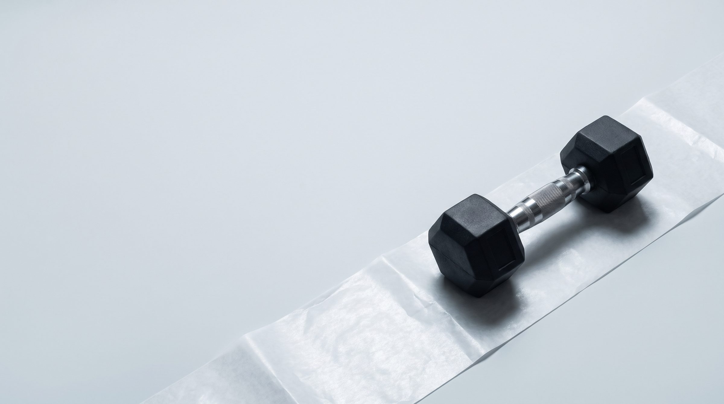 A black dumbbell resting on a strip of white exam-table paper in a pale grey studio